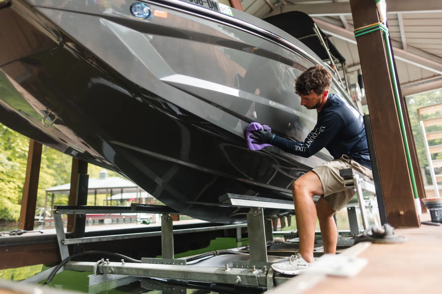 Man in shorts and gloves cleans the side of a black boat at a dock with a purple cloth.