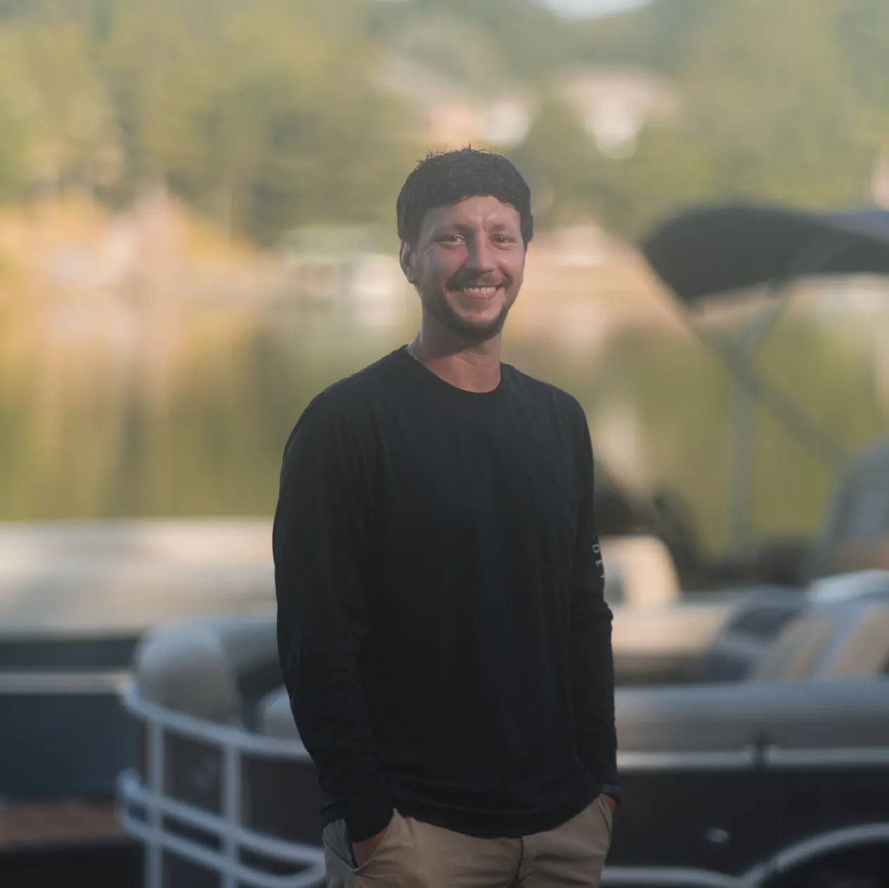 Man in a black long-sleeve shirt smiles outdoors by a lake with blurred boats in the background.