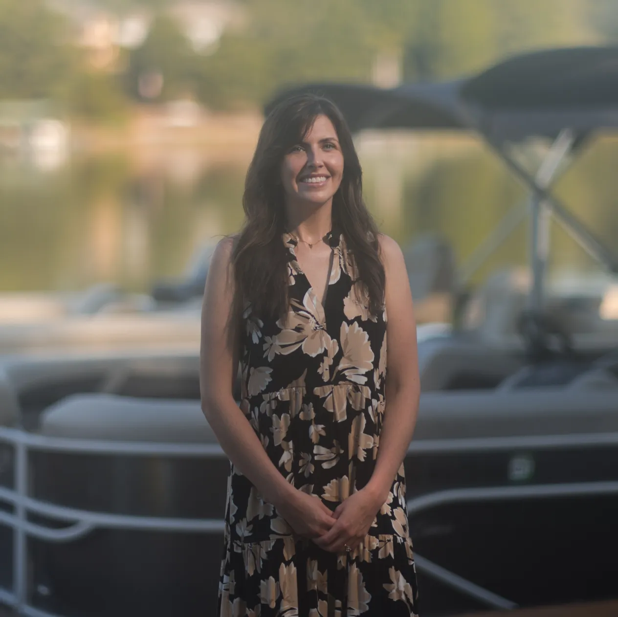 Woman in a black floral dress standing and smiling in front of a blurred boat and water background.