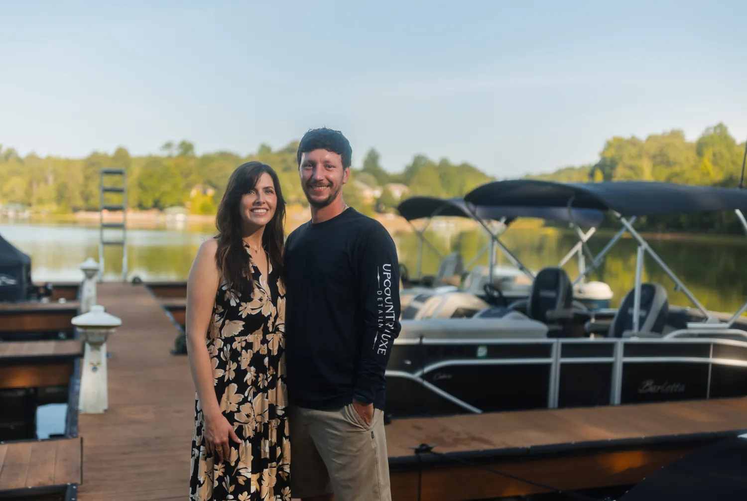 A smiling couple stands on a dock by pontoon boats, with trees and houses in the background.