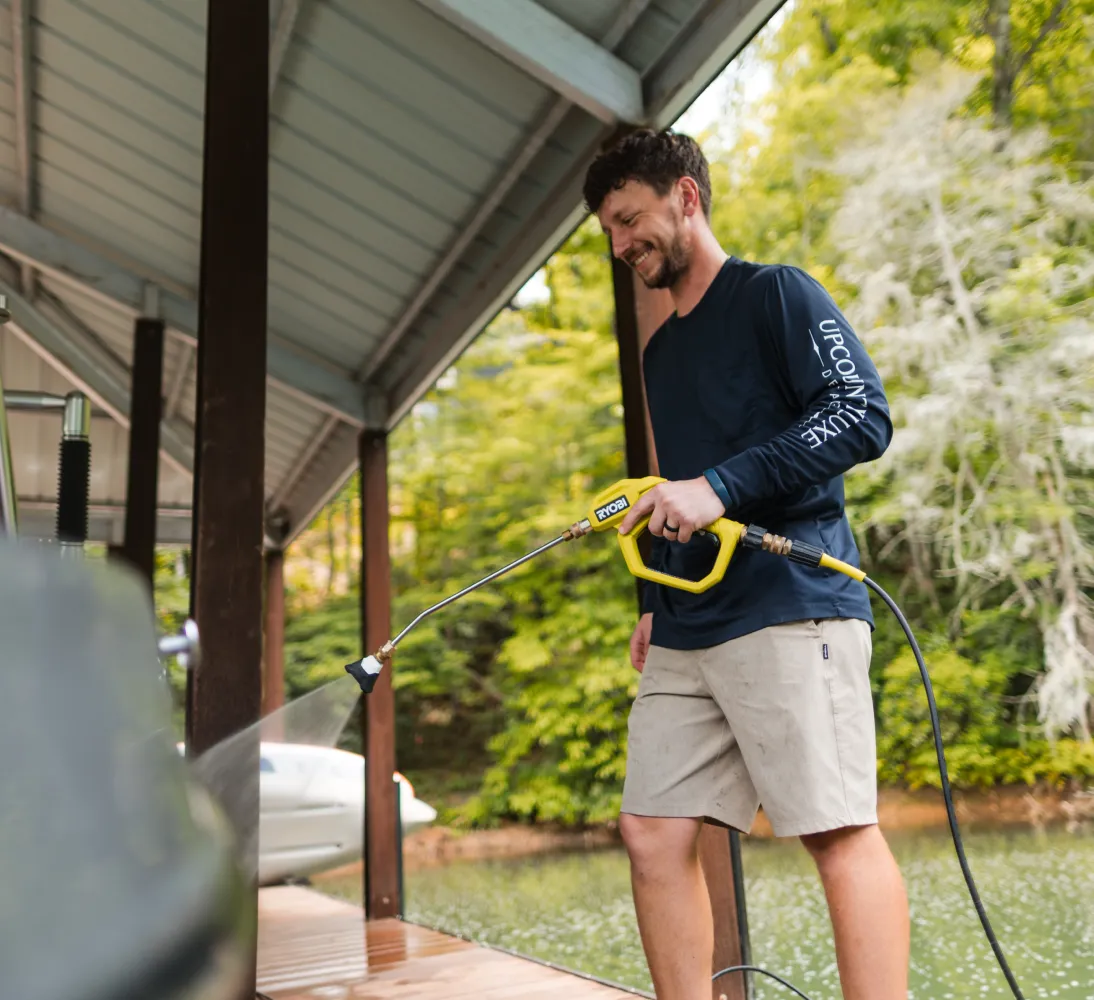 Man in shorts pressure washes a dock by the water, surrounded by trees, smiling while he works.