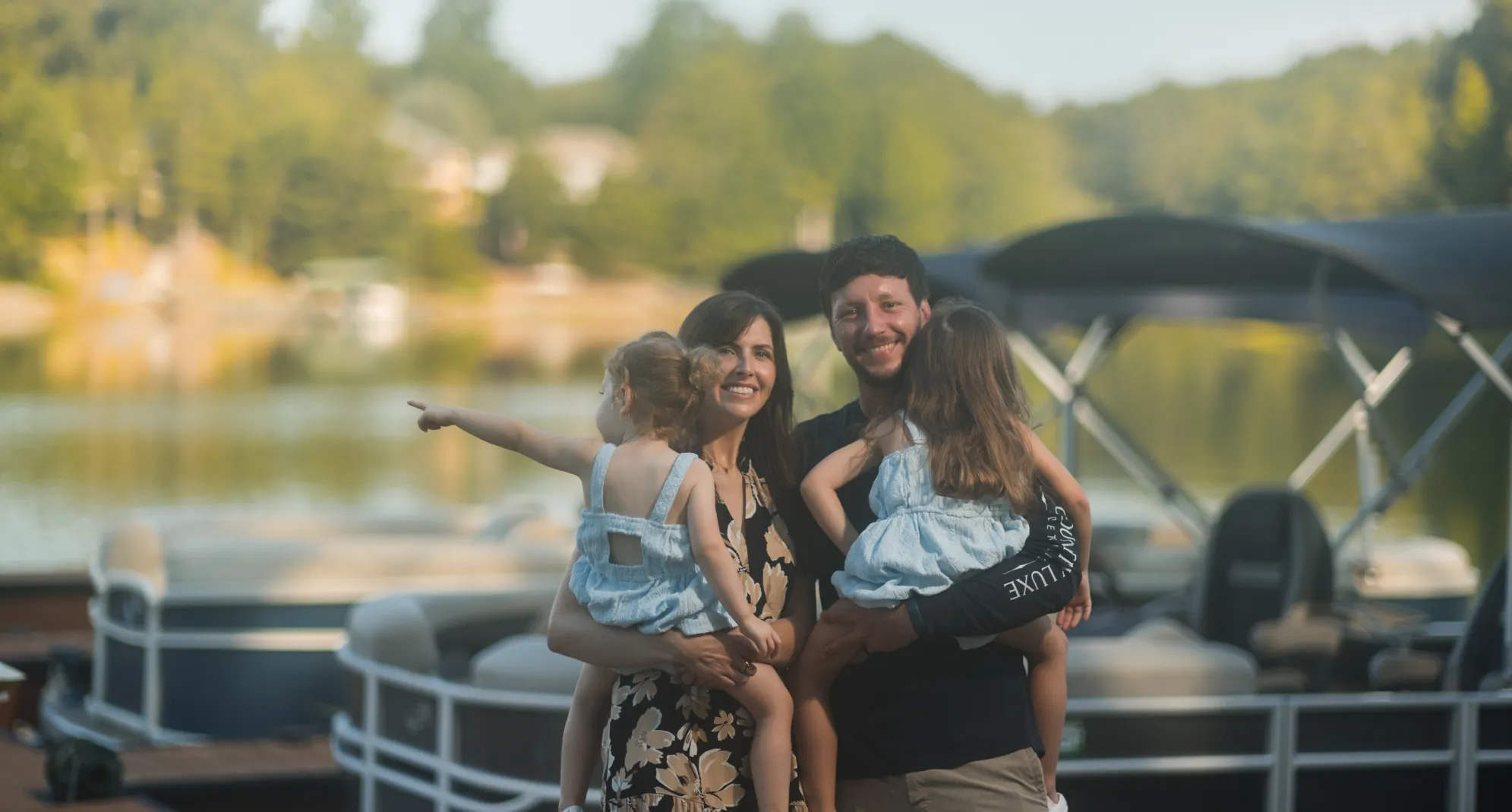 A smiling couple holds two young girls near boats on a sunny lakeside, trees in the background.