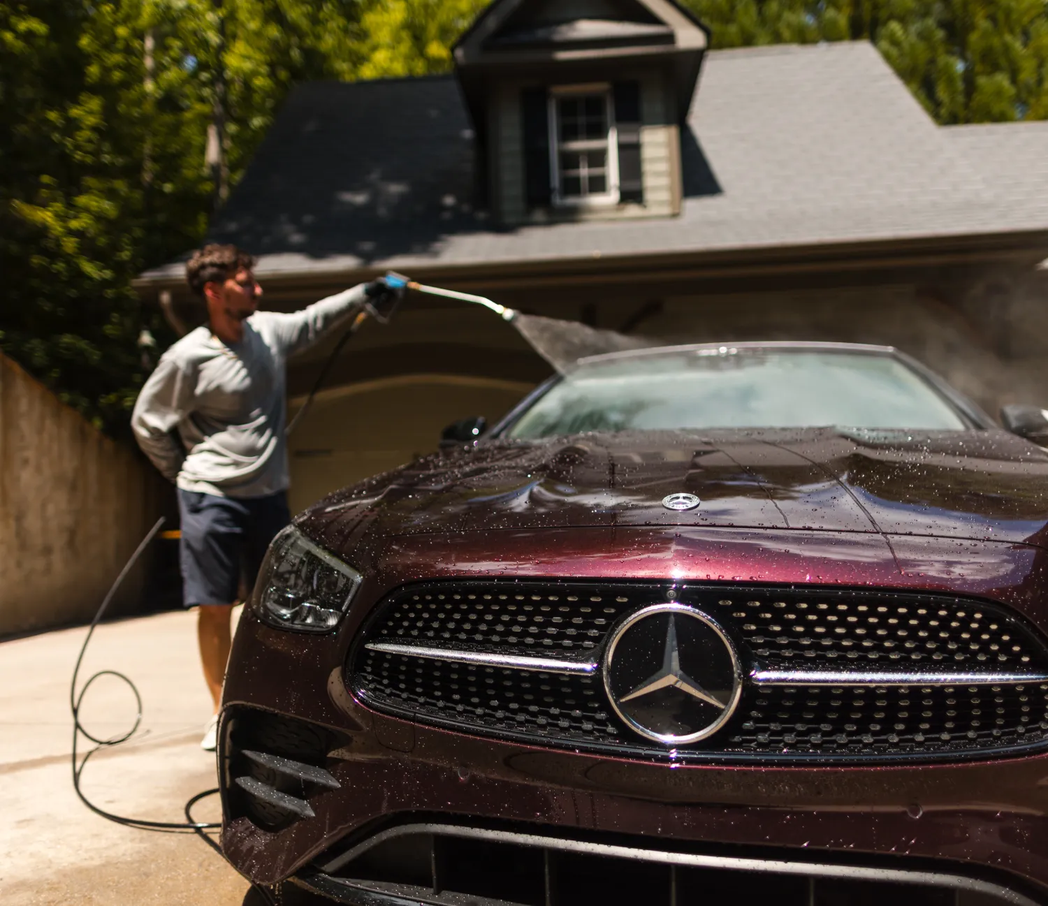 A person washes a dark Mercedes-Benz car with a hose in a driveway, house in the background.