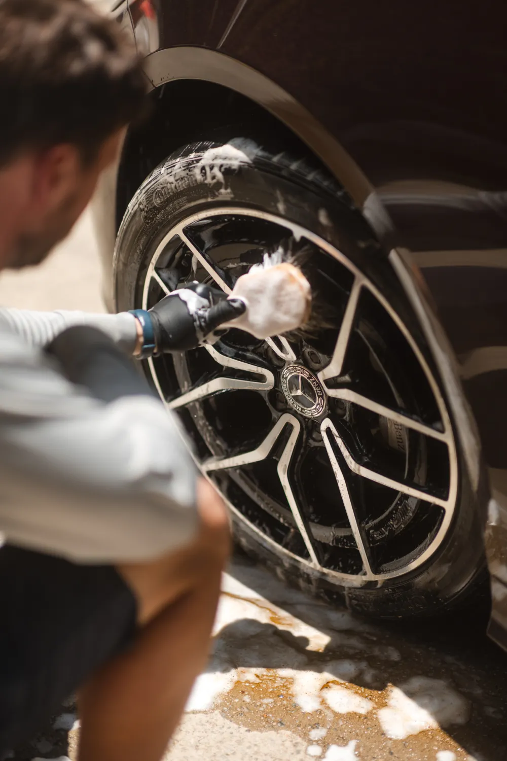 Person cleaning a black car wheel with a soapy brush, focusing on the Mercedes-Benz rim.