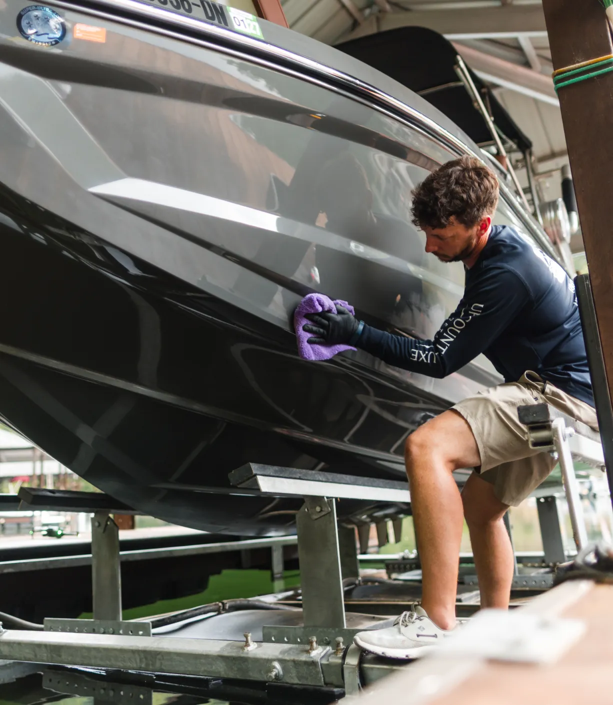 Person cleaning the side of a black boat with a purple cloth inside a covered boat storage area.