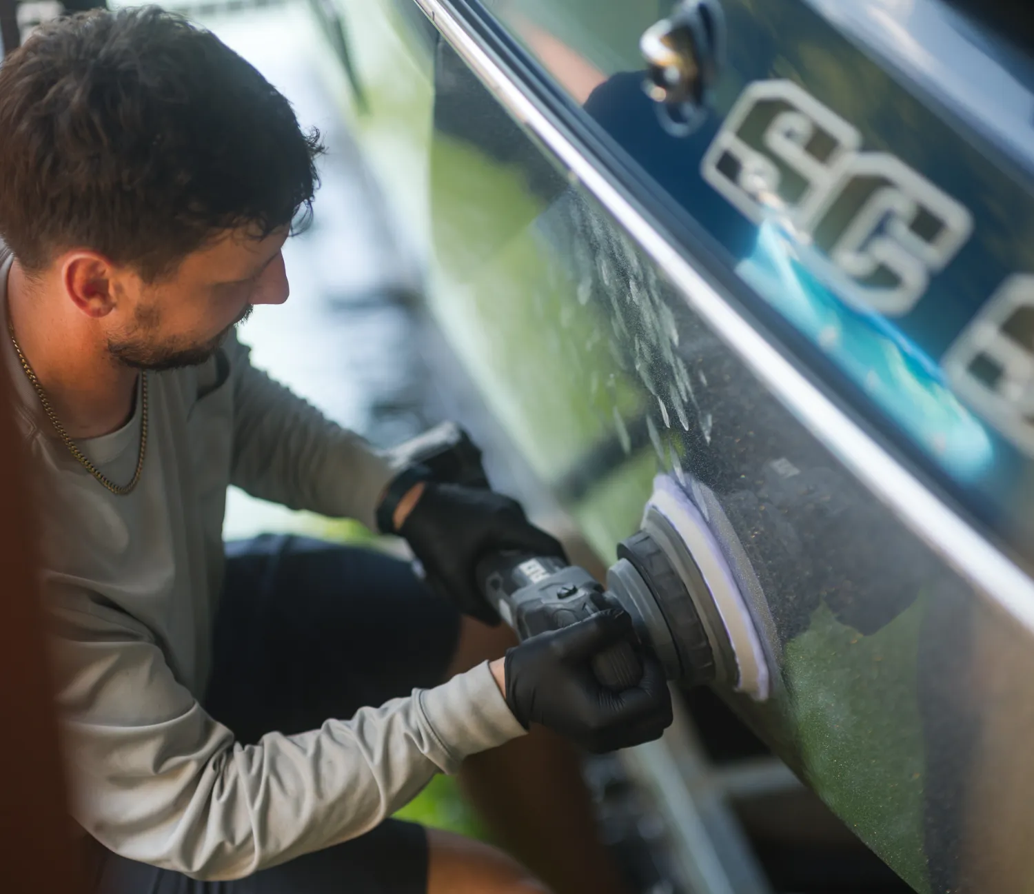 Man wearing gloves uses a power buffer to polish the side of a blue boat, reflected in the surface.