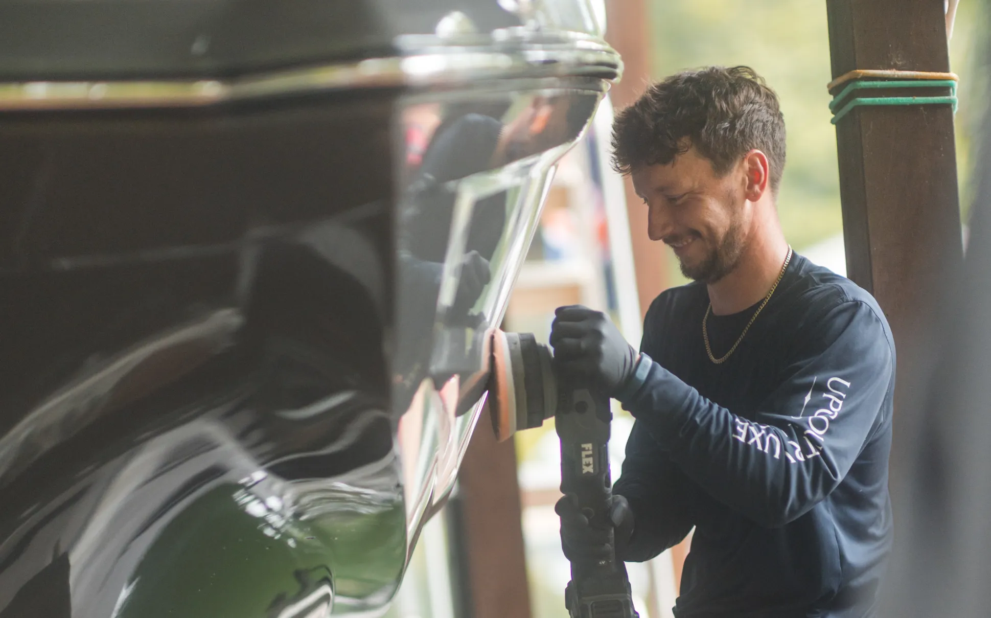 Smiling person polishes a black boat with a power buffer while wearing gloves and a long-sleeve shirt.