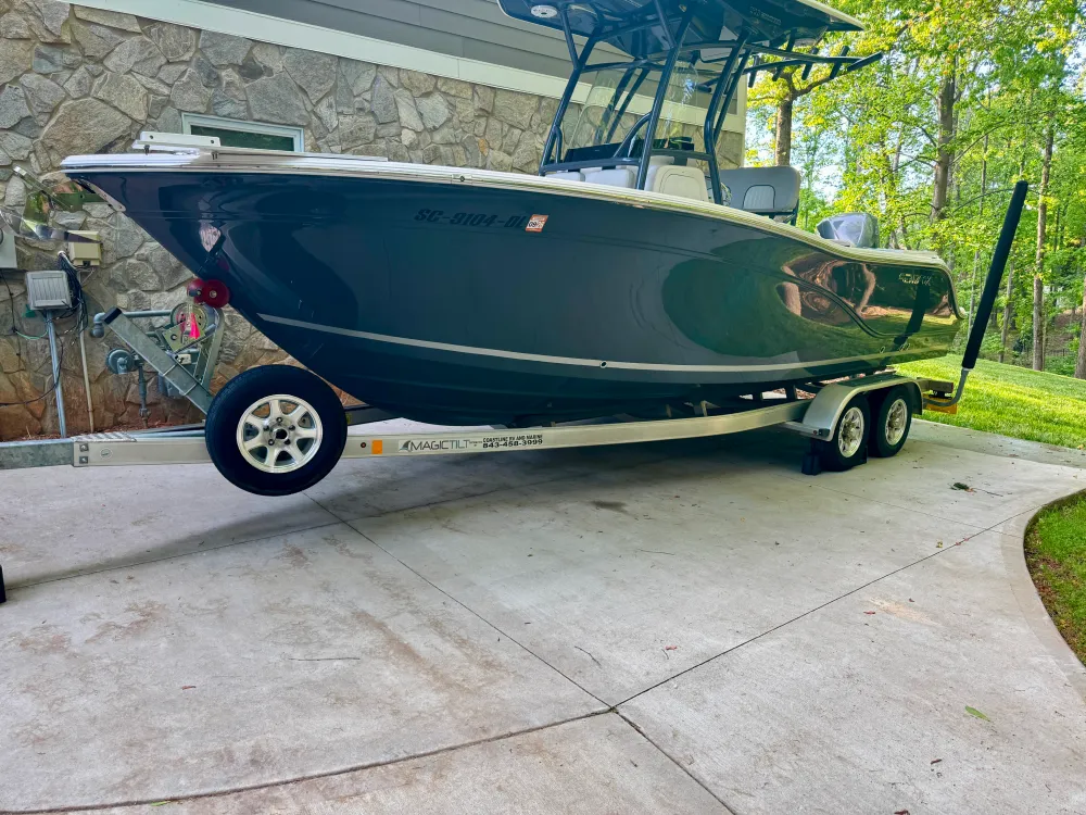 gallery-21 A black and white boat on a trailer is parked on a concrete driveway near a stone wall and green trees.