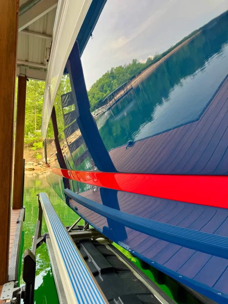 gallery-36 Docked blue boat reflects water, trees, and dock; red stripe runs along the side above the green lake.