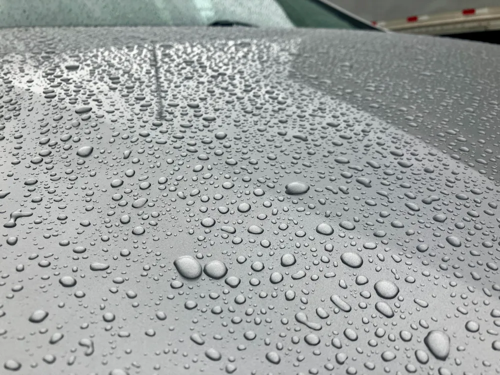 gallery-38 Water droplets scattered across a smooth gray car hood after rain, with reflections visible.