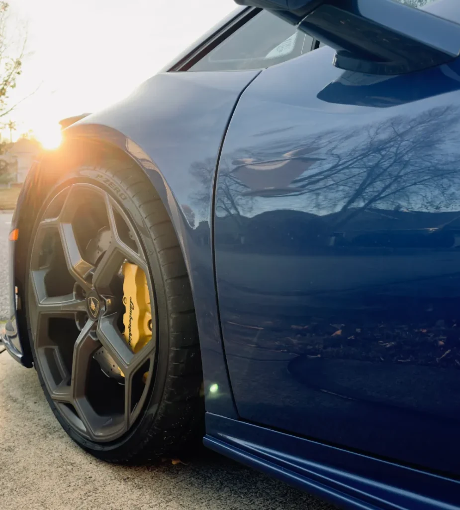 gallery-41 Close-up of a blue sports car’s front wheel with yellow brake caliper, sunlight shining on the side.