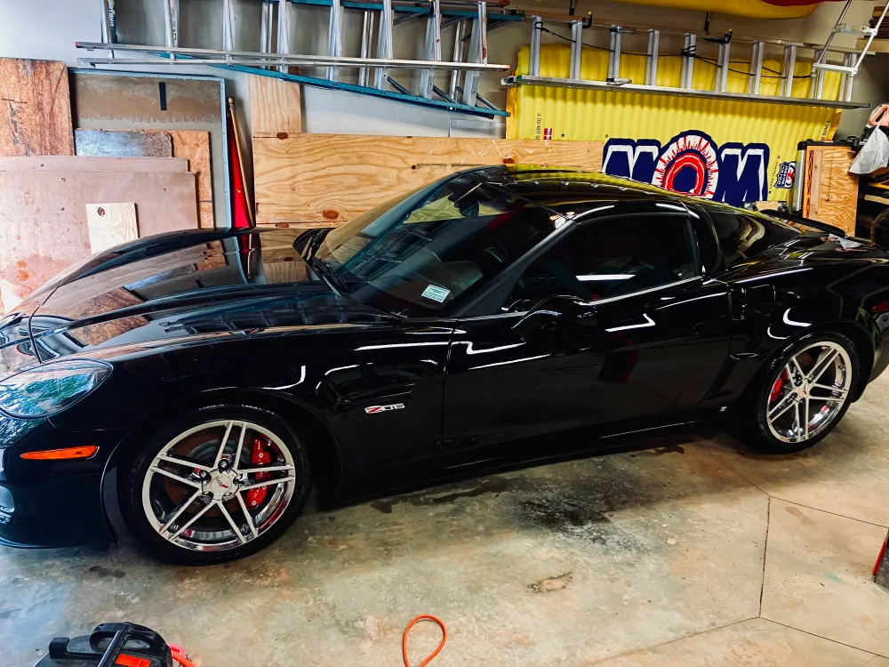 gallery-43 Black Chevrolet Corvette Z06 parked in a garage with tools, wood panels, and a yellow sign in the background.