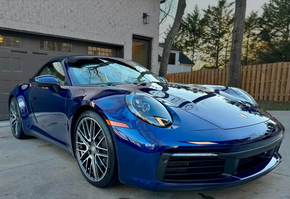 gallery-44 Blue Porsche sports car parked on driveway in front of a garage, with trees and a fence in the background.