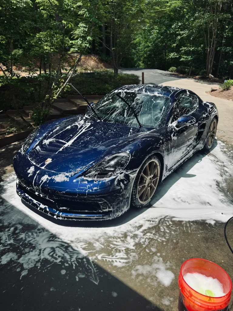 gallery-45 A blue sports car covered in soap suds is being washed outside on a sunny day, with a red bucket nearby.