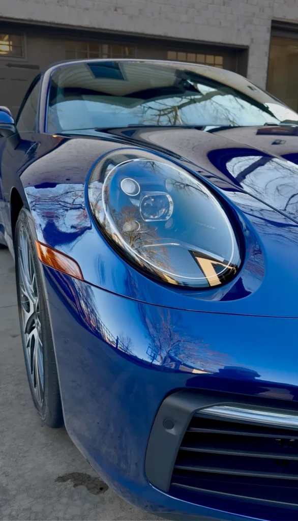 gallery-46 Close-up of a shiny blue sports car’s front left headlight, with trees reflected on the hood.