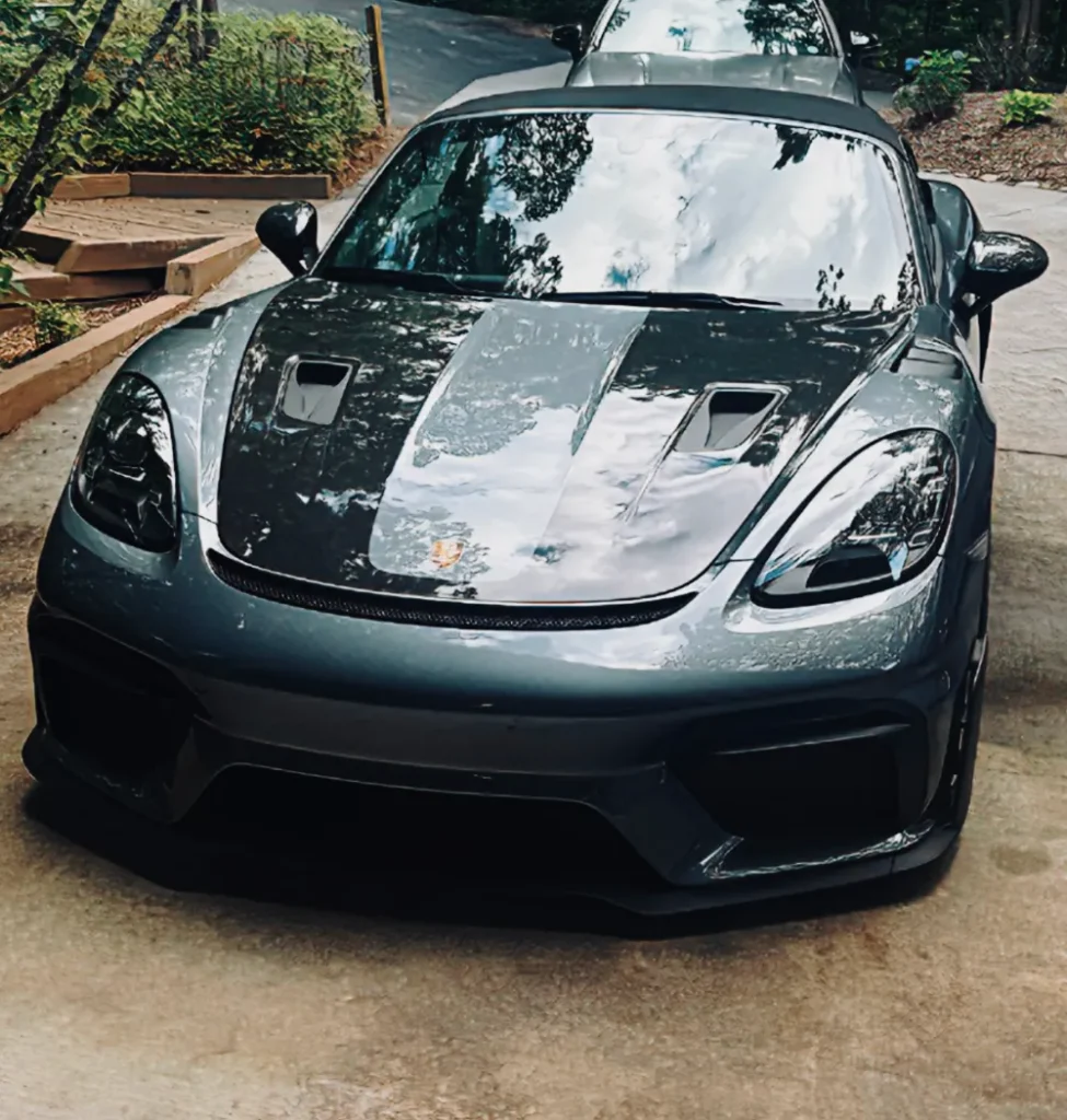 gallery-50 A dark gray Porsche sports car is parked on a driveway, with trees and sky reflected on its hood.