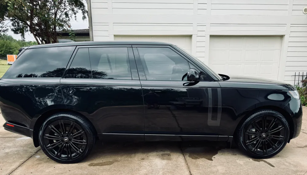 gallery-53 Black SUV parked on a driveway in front of a white garage, with trees and grass visible in the background.