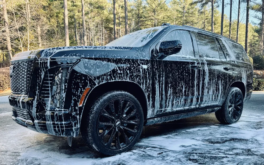 gallery-7 Black SUV covered in white soap suds parked outdoors, surrounded by trees in the background.