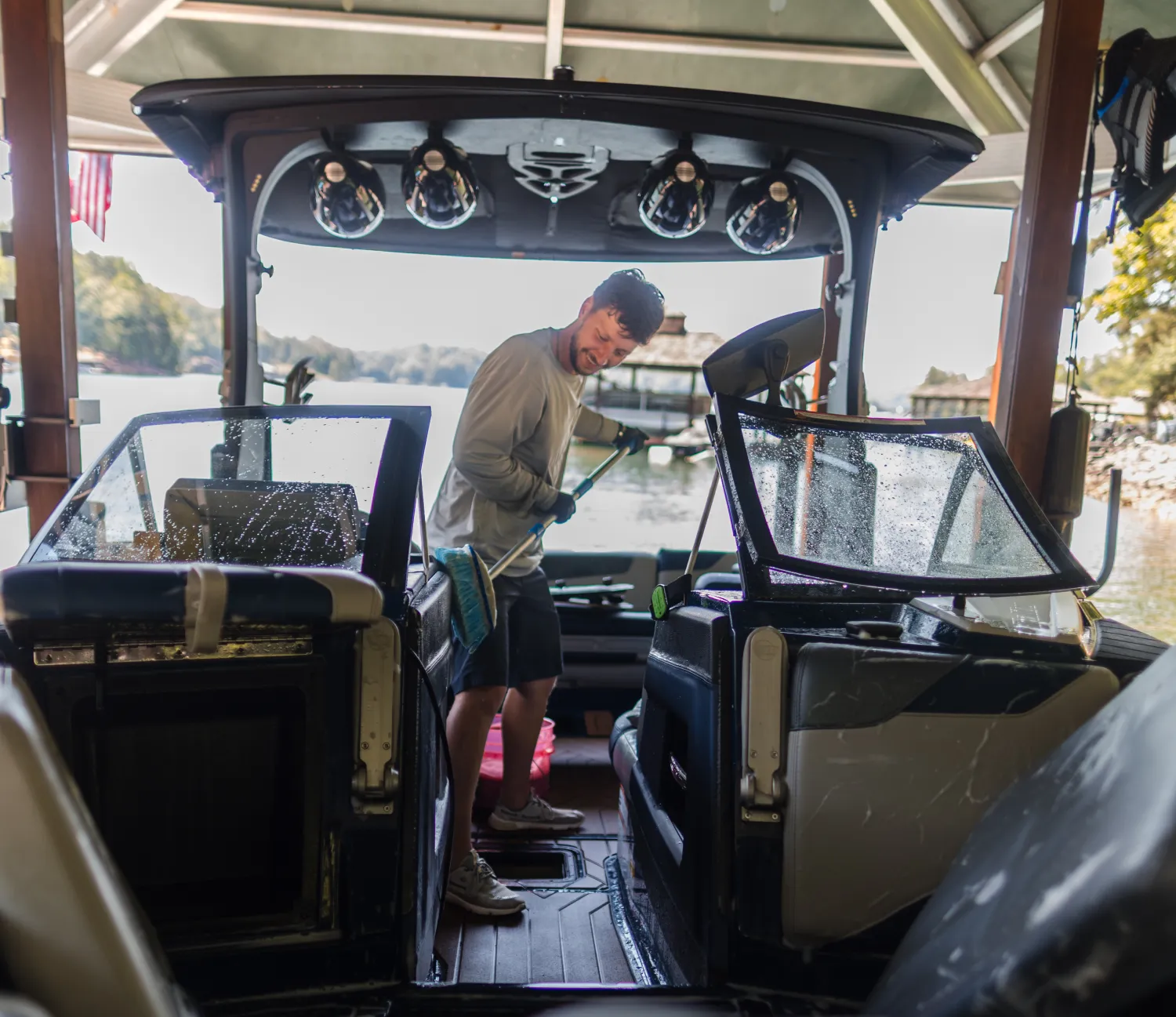 Man cleaning the interior of a boat with a broom, dock and water visible in the background.