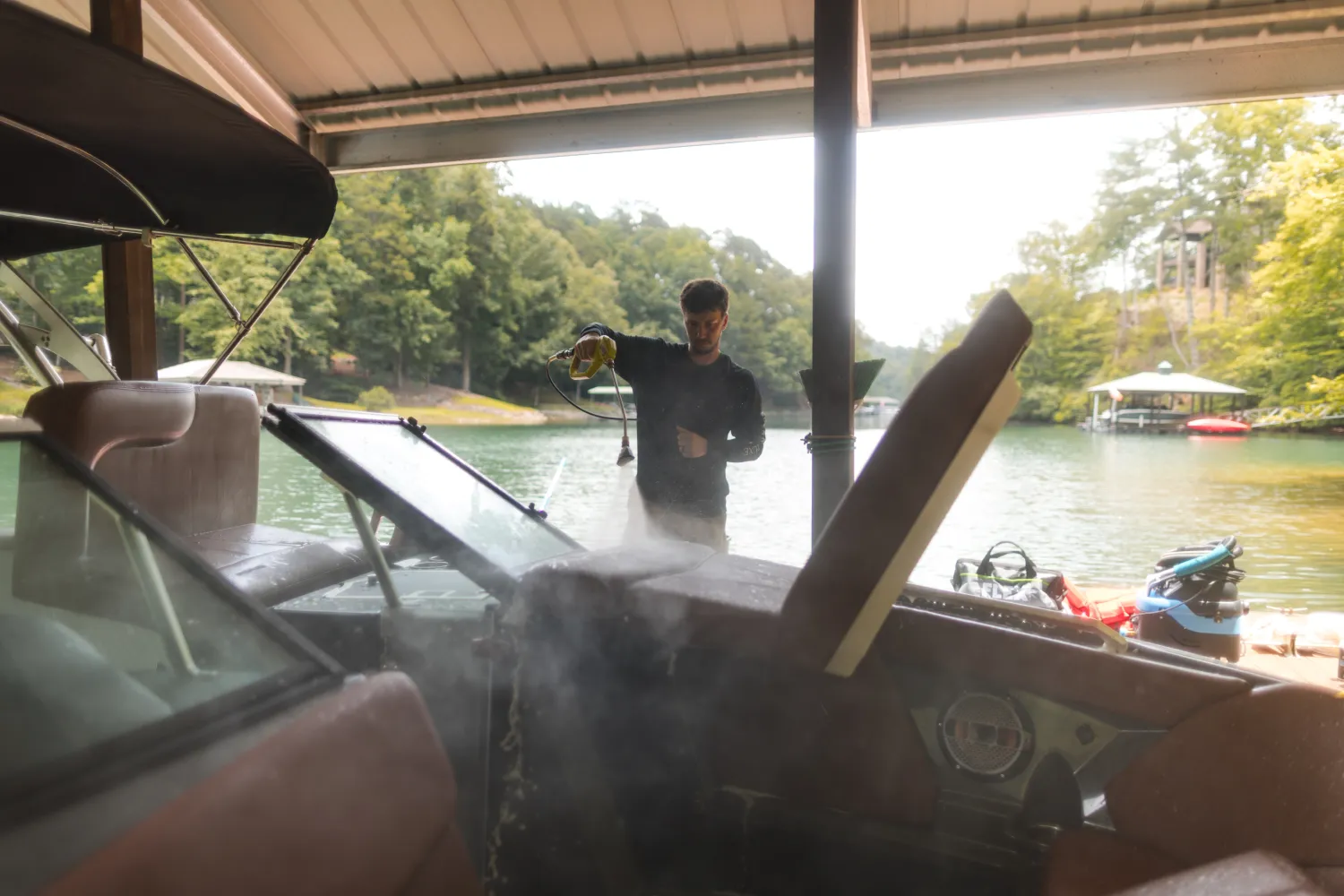 A person sprays water to clean a boat at a dock near a lake, with trees in the background.