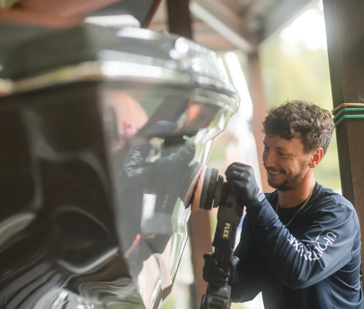 Man smiling while polishing a boat’s shiny surface with a power tool under a covered area.