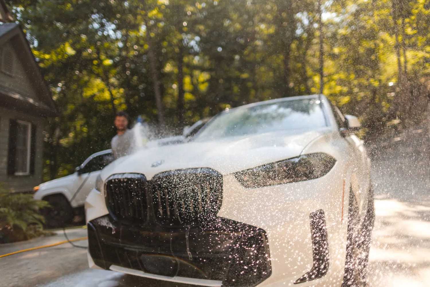 A white BMW is being washed outdoors, water spraying on its front grille with trees in the background.
