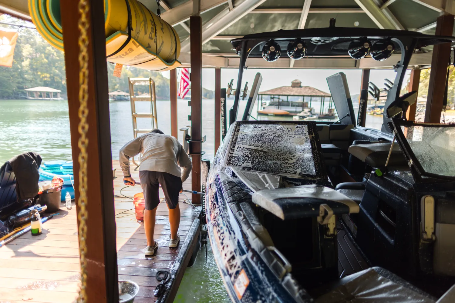 Person cleaning a boat at a lakeside dock under a roof, with sunlight reflecting on the water nearby.