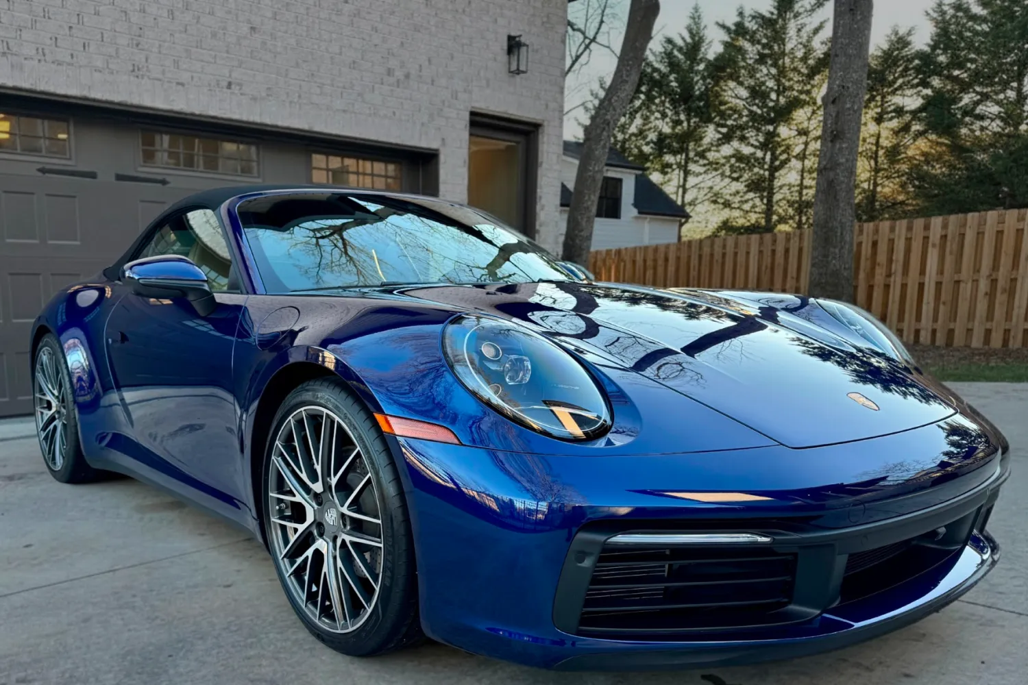 A blue Porsche sports car parked in a driveway near a house with a garage and wooden fence.