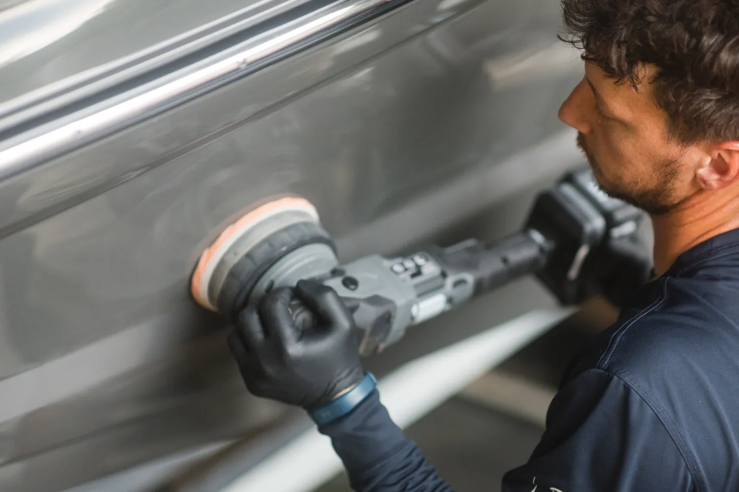 Person polishing a gray boat’s surface with a power buffer while wearing black gloves.