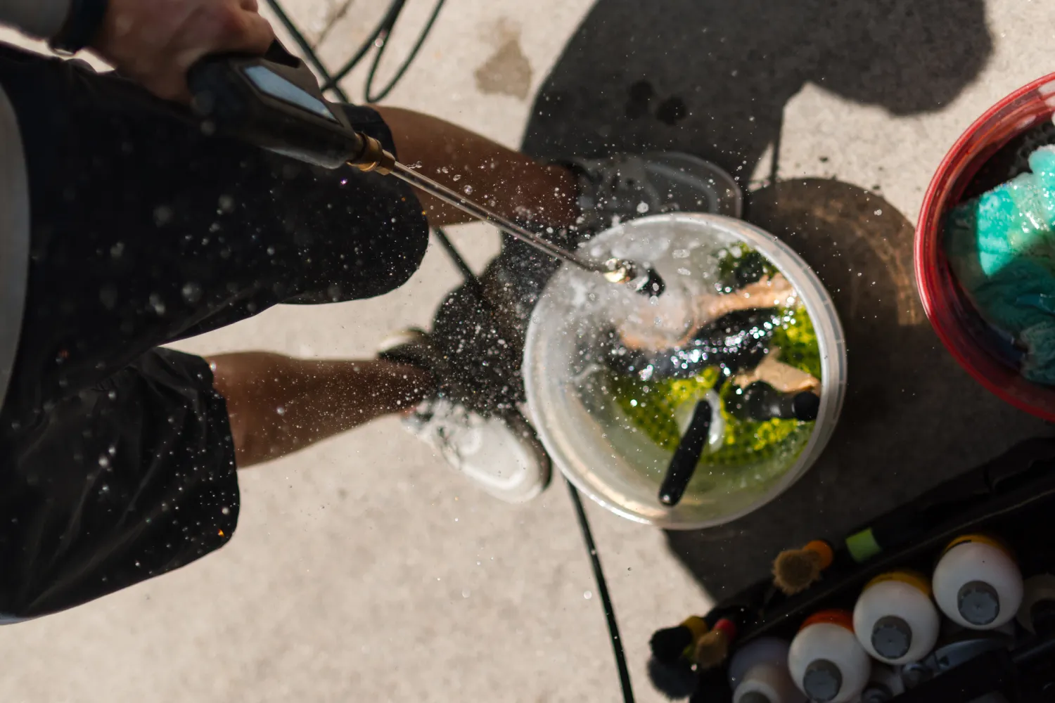 Person rinsing black tools in a bucket with a hose, with water splashing and cleaning supplies nearby.