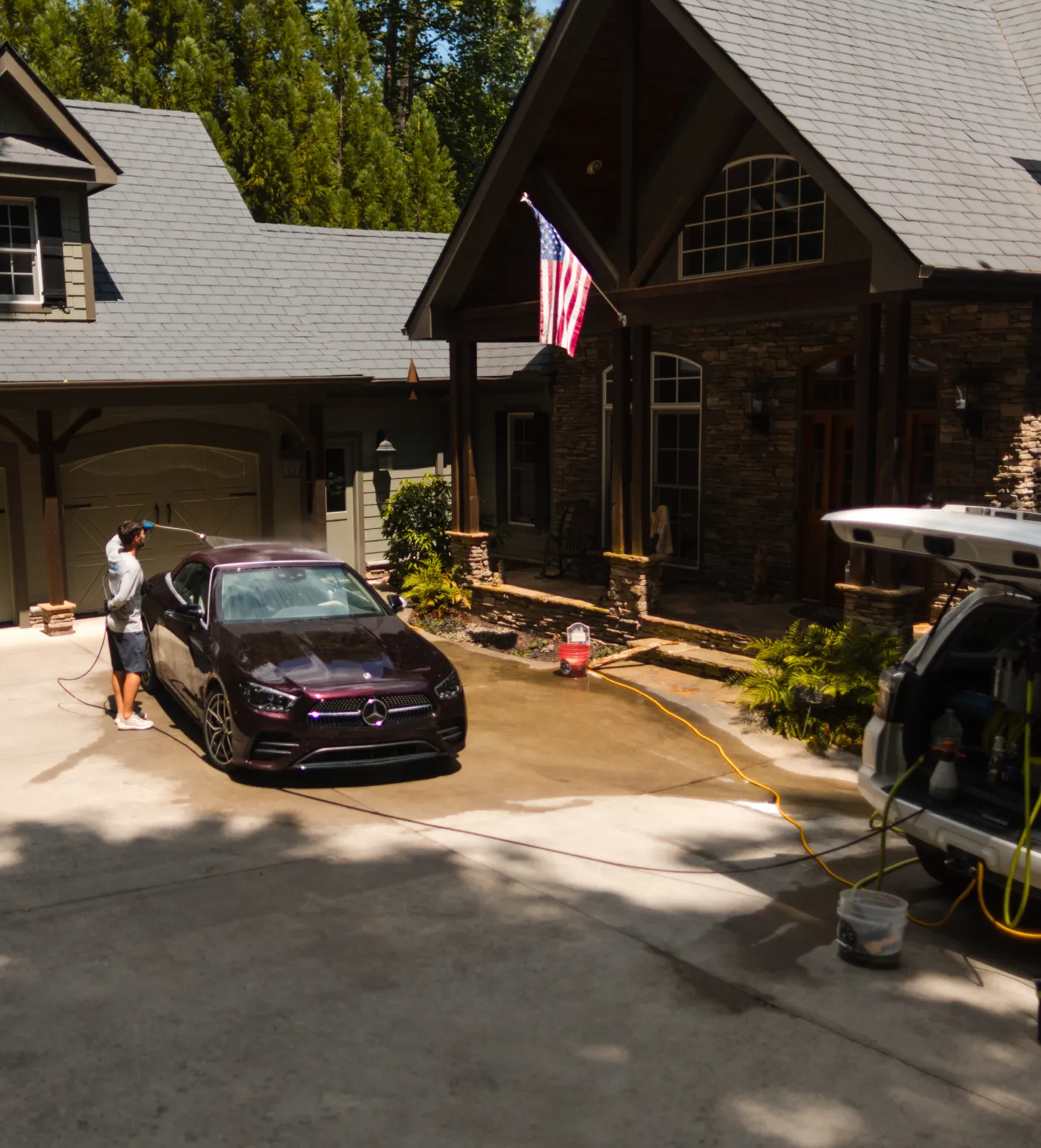 Person washing a black Mercedes in a driveway outside a large house with an American flag.