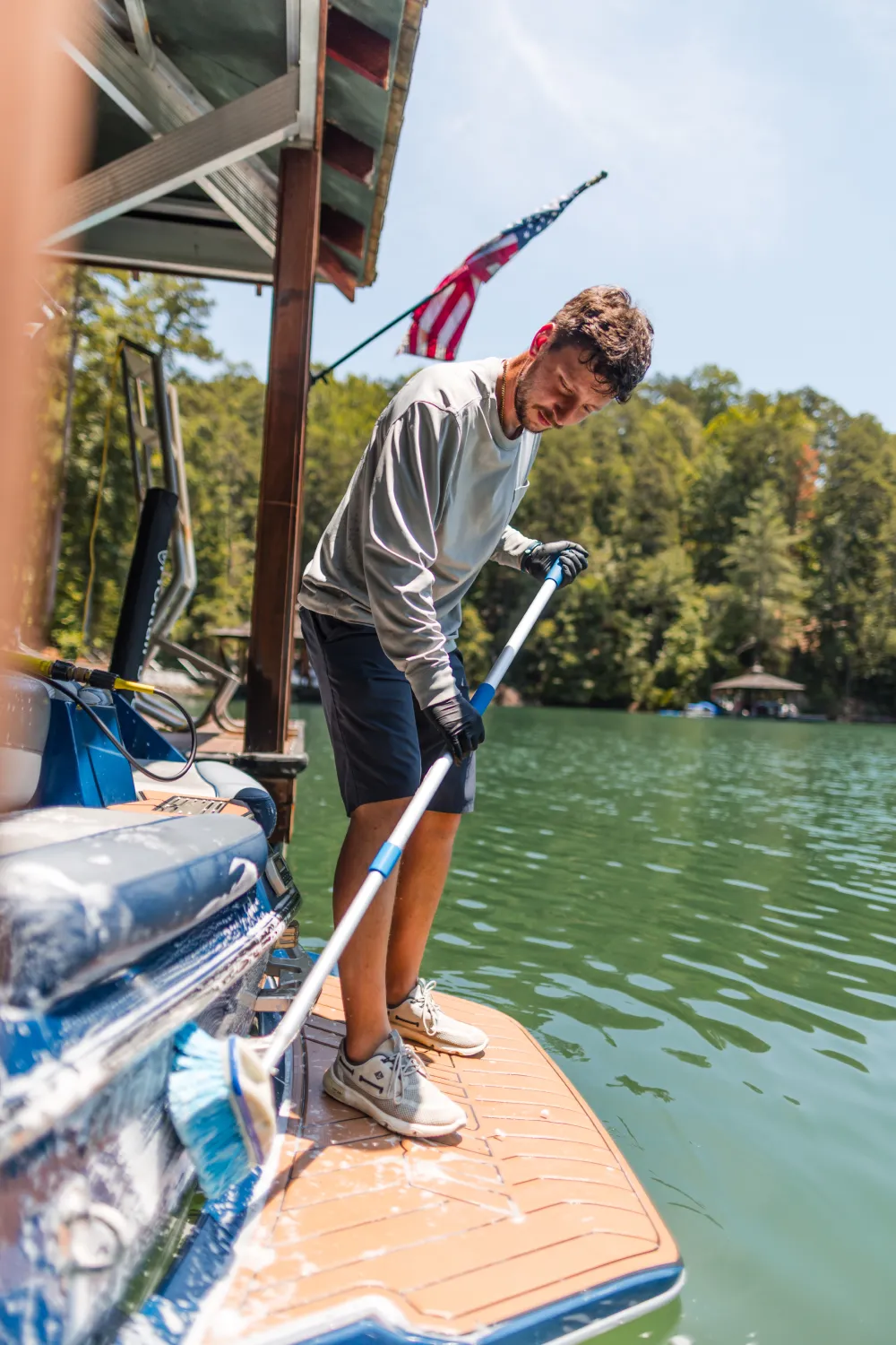 Man in gloves cleaning a boat dock with a brush on a sunny day; water and trees in background.